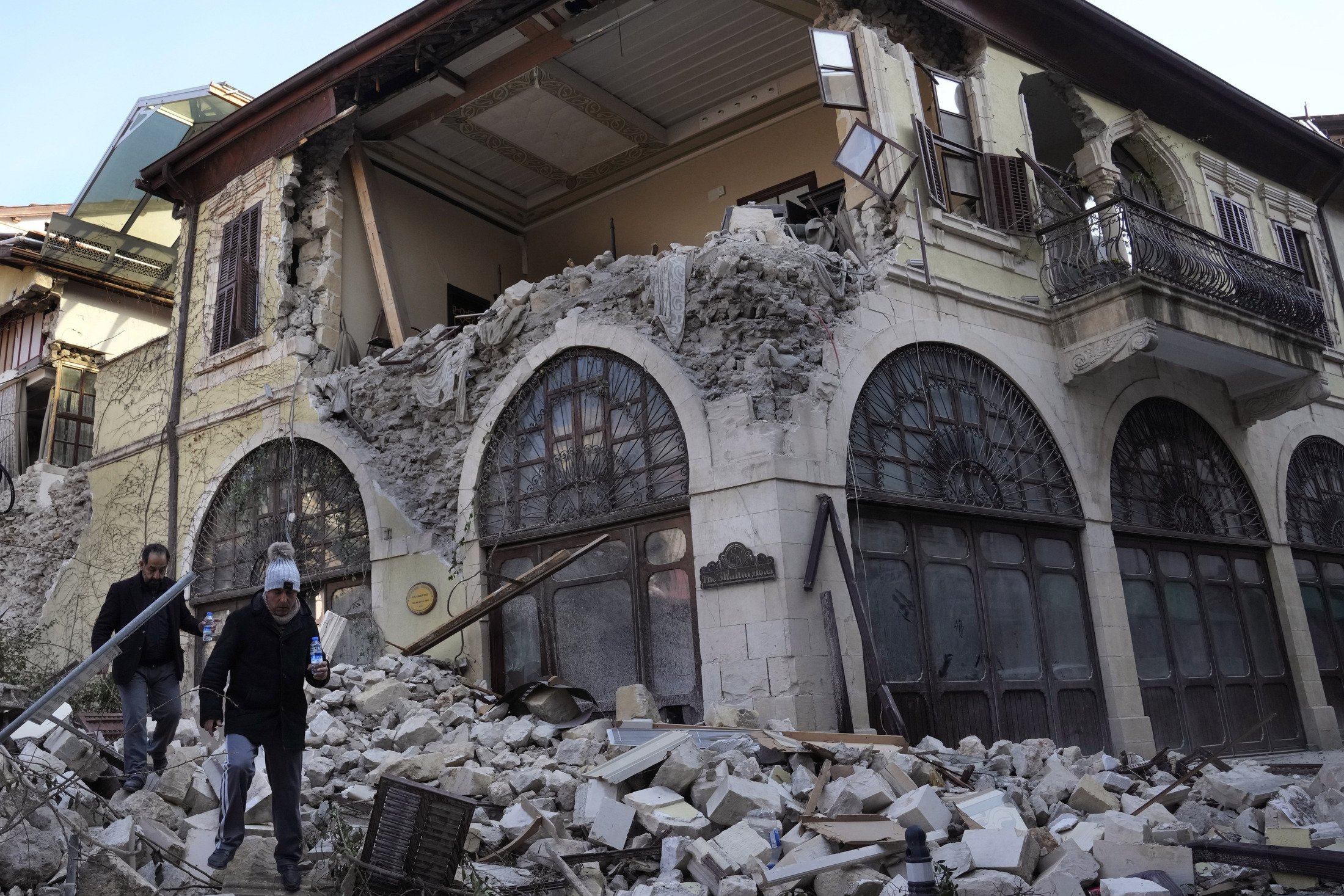 Türk vatandaşları, 13 Şubat 2023, Antakya'nın eski kentinde, büyük depremde yıkılan tarihi bir otelin önünden geçiyor. (AP Fotoğrafı)