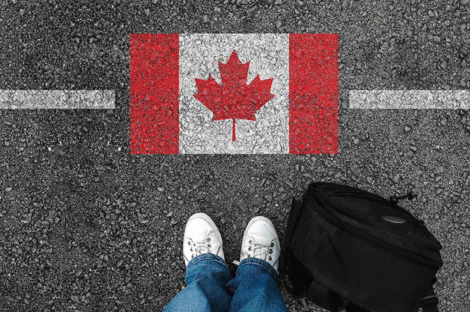 A man stands on the asphalt next to a flag of Canada near the country&#039;s border with the U.S.