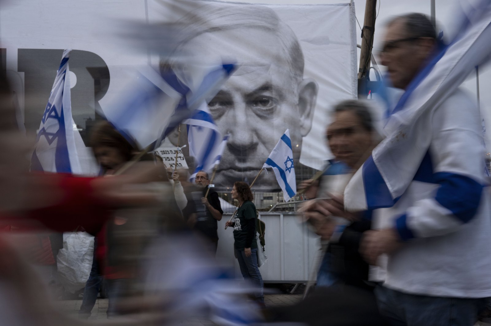 Demonstrators walk with Israel&#039;s national flags next to a banner showing Prime Minister Benjamin Netanyahu during a protest against plans by his government to overhaul the judicial system in Tel Aviv, Israel, March 25, 2023. (AP Photo)