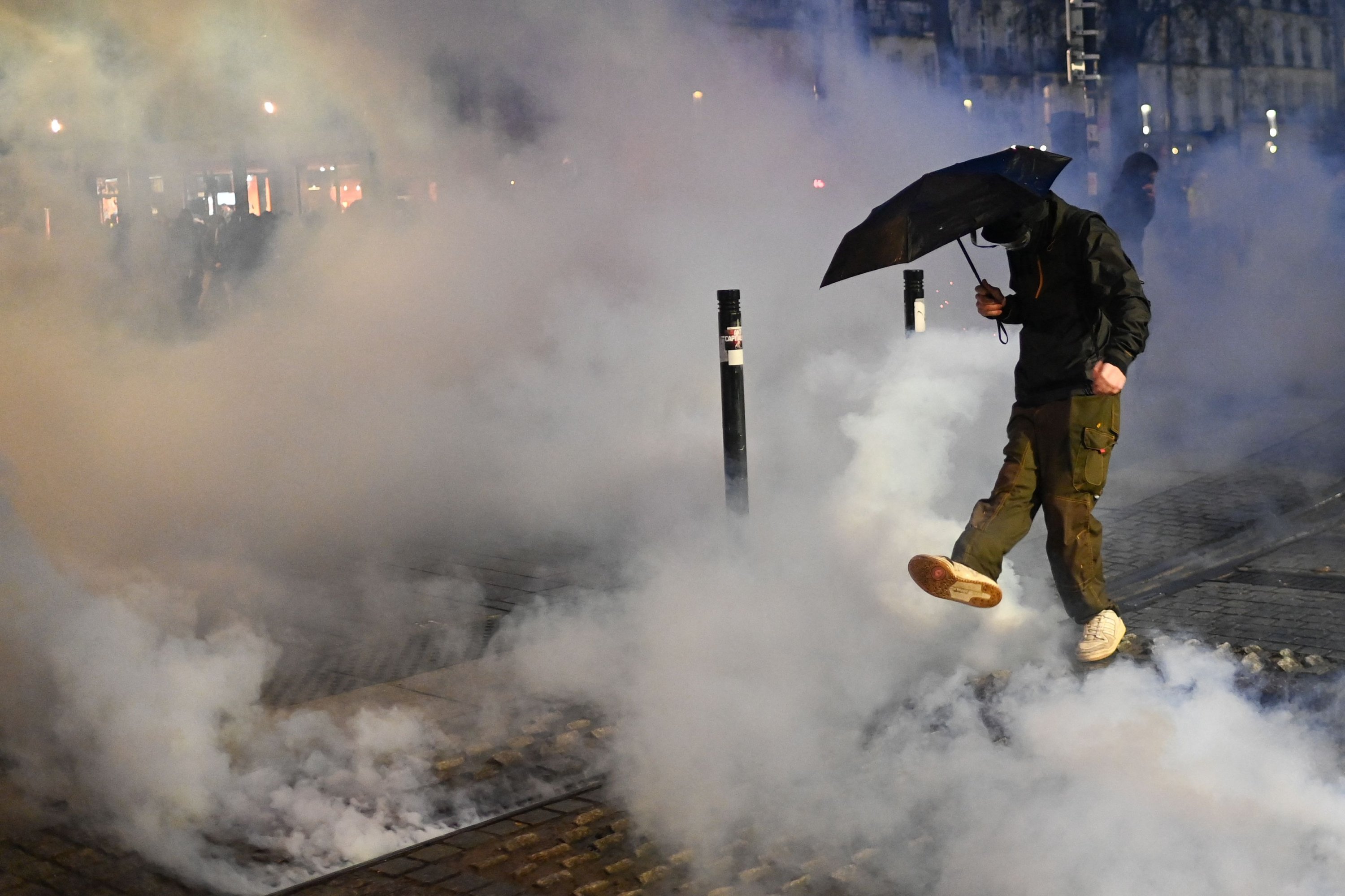 Seorang pengunjuk rasa yang memegang payung berusaha menendang tabung gas air mata yang berasap selama demonstrasi untuk mendukung para korban kebrutalan polisi, setelah peristiwa di Sainte-Soline dan di tengah protes pensiun, di Nantes, Prancis barat, 30 Maret 2023. (Foto AFP )