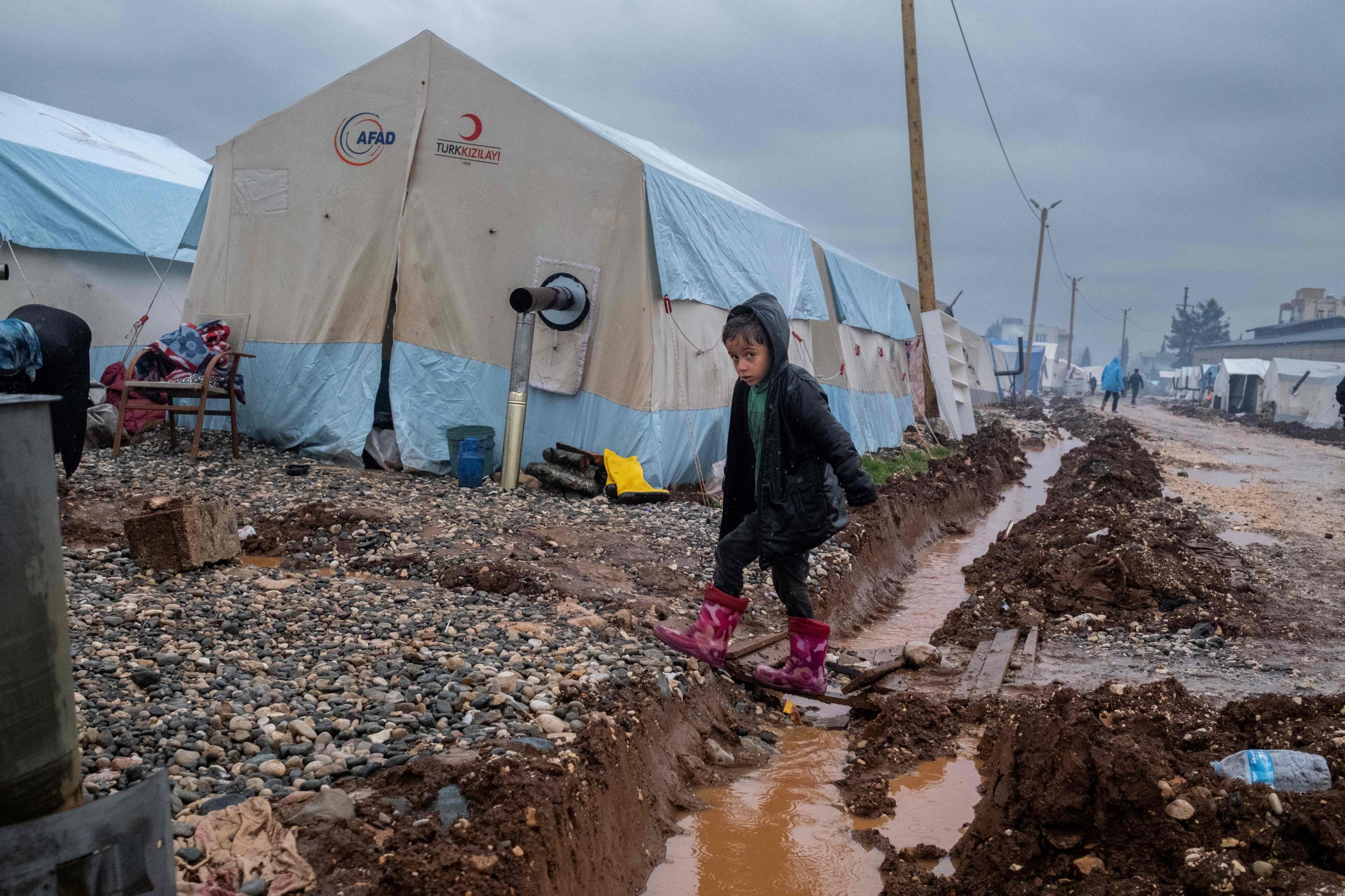 Seorang anak melintasi selokan air di bawah hujan untuk mencapai tenda yang didirikan untuk menampung para pengungsi setelah gempa besar bulan lalu, di Adıyaman, Türkiye tenggara, 25 Maret 2023. (Foto AFP)