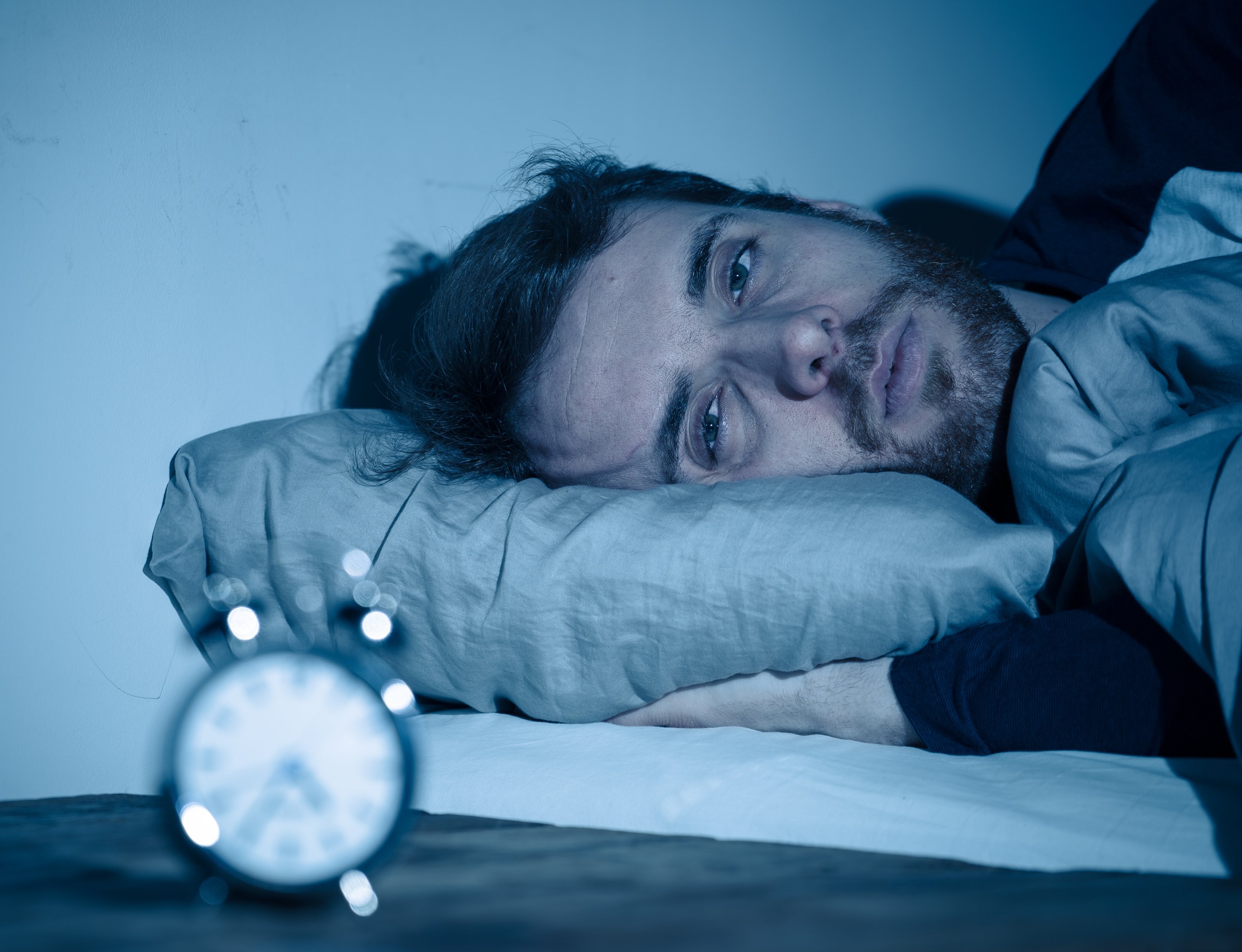 Young man awake at night not able to sleep, feeling frustrated and worried looking at the clock. (Shutterstock Photo)