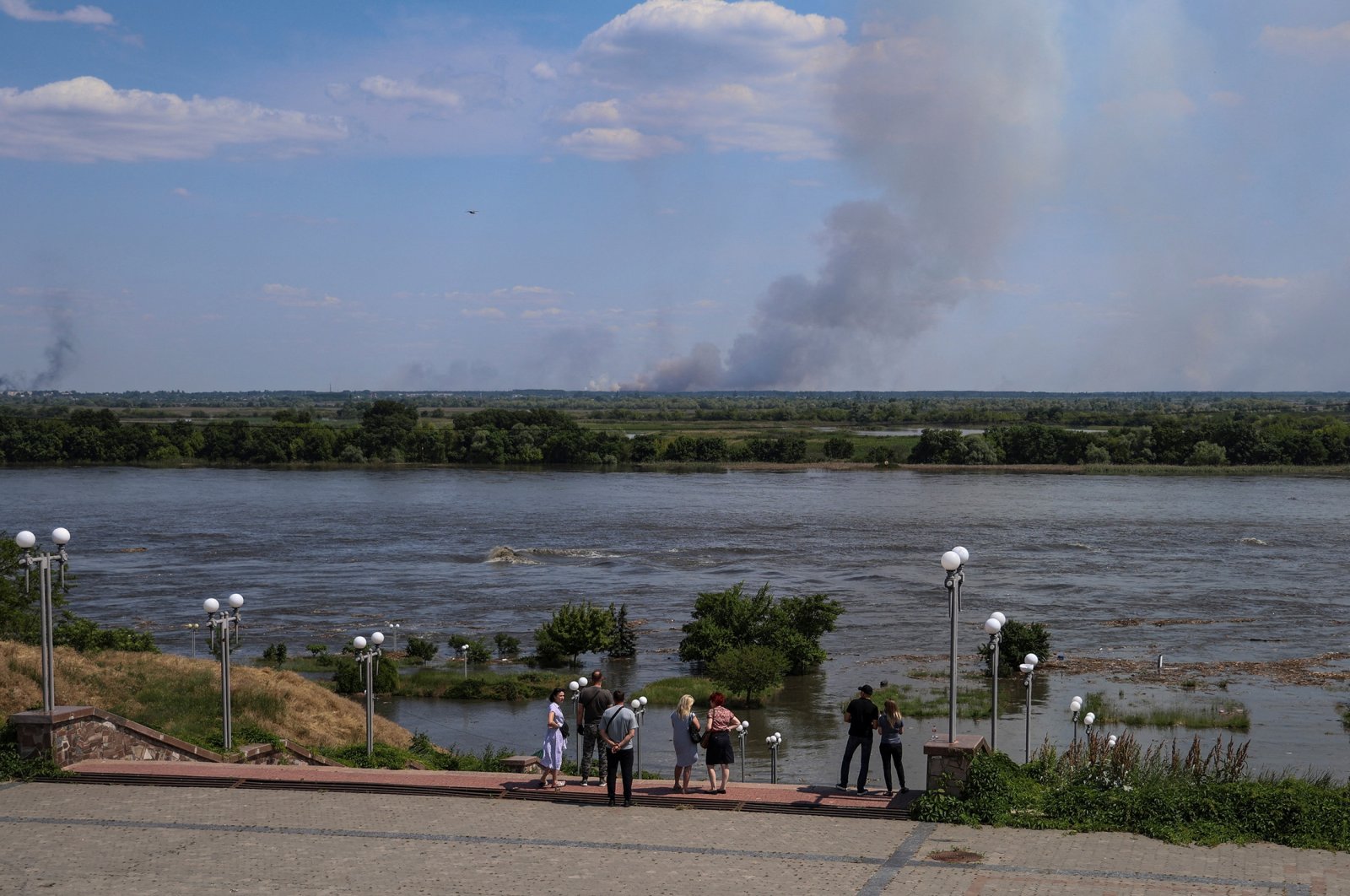 Local residents stand on an embankment of the Dnipro river which flooded after the Nova Kakhovka dam breached as smoke billows after shelling on the opposite bank, amid Russia&#039;s attack on Ukraine, in Kherson, Ukraine, June 6, 2023. (Reuters Photo)