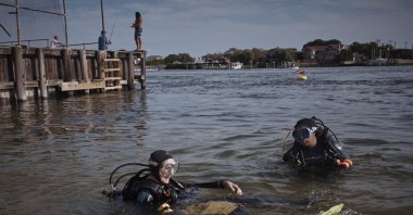 A man fishes, top second left, as scuba divers, Sarah Sears, bottom left, and Tanasia Swift, (L), prepare to enter the water during an underwater cleanup in the Queens borough of the U.S., Aug. 27, 2023. (AP Photo)