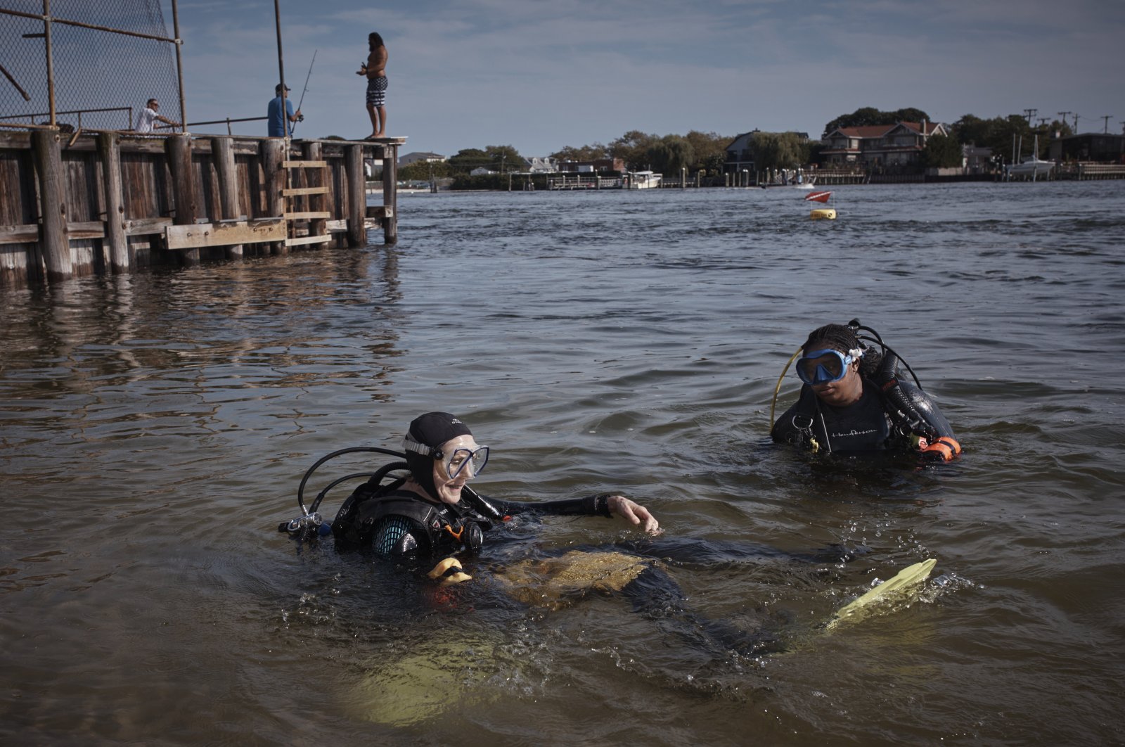 A man fishes, top second left, as scuba divers, Sarah Sears, bottom left, and Tanasia Swift, (L), prepare to enter the water during an underwater cleanup in the Queens borough of the U.S., Aug. 27, 2023. (AP Photo)