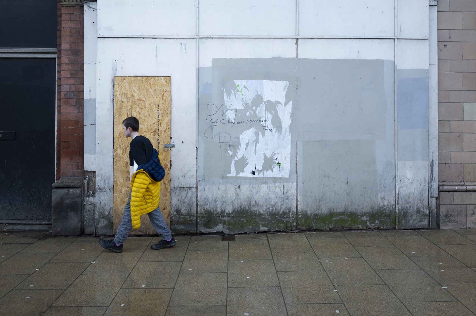 In rainy weather, a young boy in a yellow coat walks past a plyboarded and graffiti-covered retail space near the high street in Leeds, United Kingdom, Feb. 24, 2022. (Getty Images Photo)