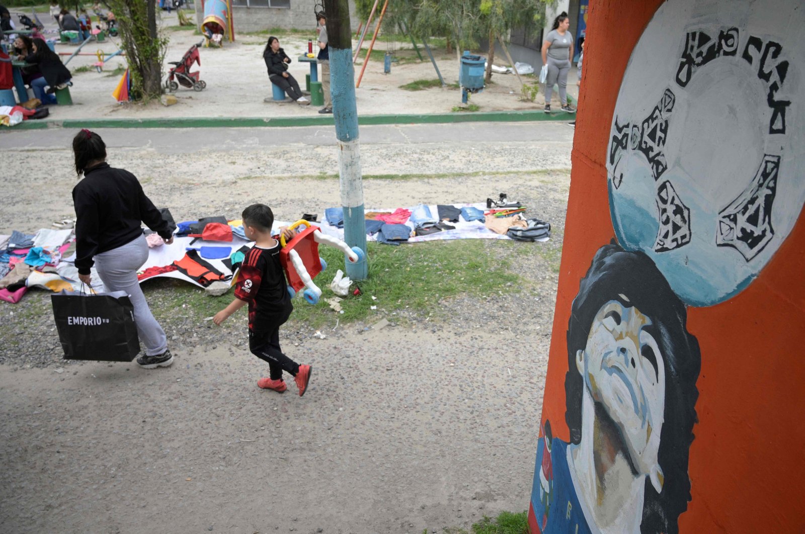 People pass by used clothes for barter or sale on a sidewalk in Villa Fiorito, Lomas de Zamora where a pedestrian bridge is decorated with portraits of late Argentine football star Diego Maradona, Argentina, Nov. 13, 2023. (AFP Photo)