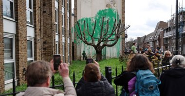 Crowds gather to view a Banksy artwork, a stencil of a person having spray painted tree foliage onto a wall behind a leafless tree, a graffiti artwork confirmed as being the work of the famous street artist near Finsbury Park in north London, U.K., March 18, 2024. (AFP Photo)
