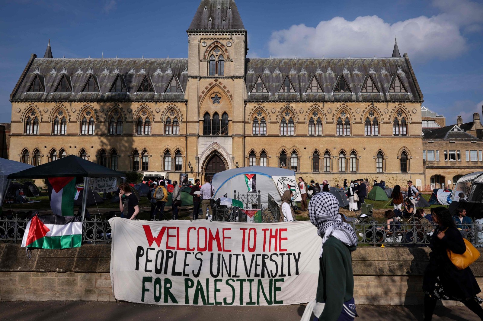 A passerby with the head covered with a keffiyeh walks past a banner reading &quot;Welcome to the people&#039;s university for Palestine&quot; at Oxford University, as a pro-Palestinian camp has been set up on the campus, in Oxford, eastern England, May 7, 2024. (AFP Photo)