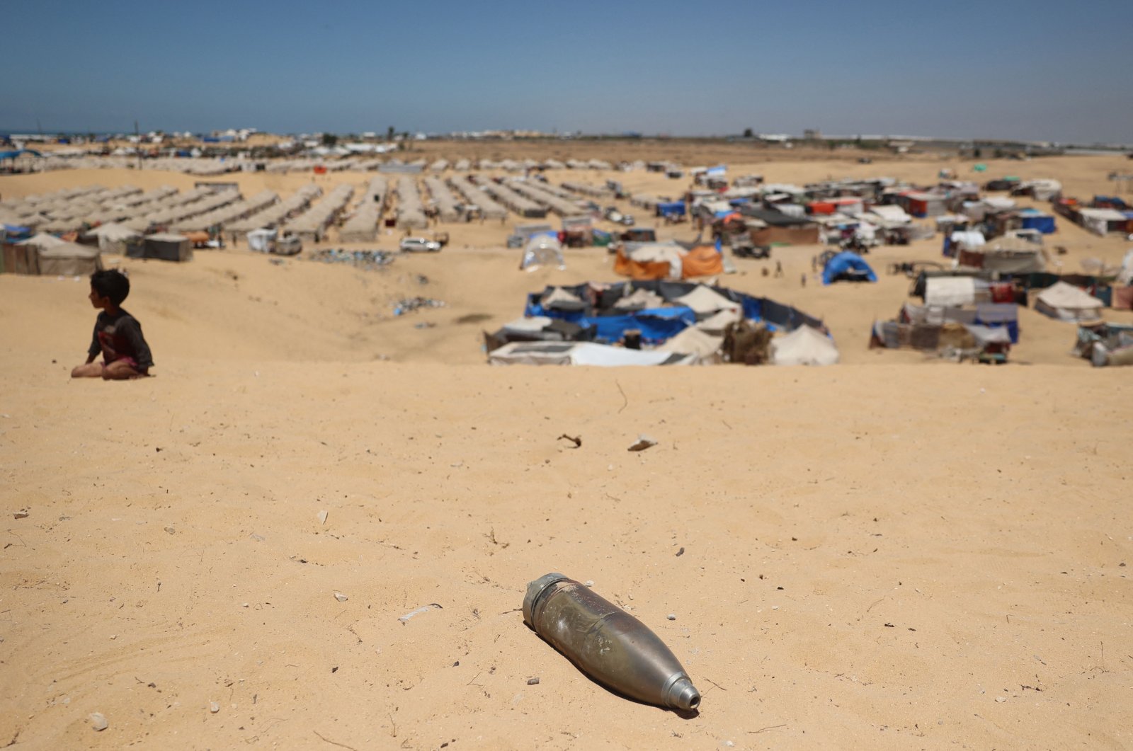 An unexploded Israeli shell lies on a sand dune as a young boy sits near a makeshift camp for displaced Palestinians in the area of Tel al-Sultan, Rafah, southern Gaza Strip, Palestine, May 30, 2024. (AFP Photo)