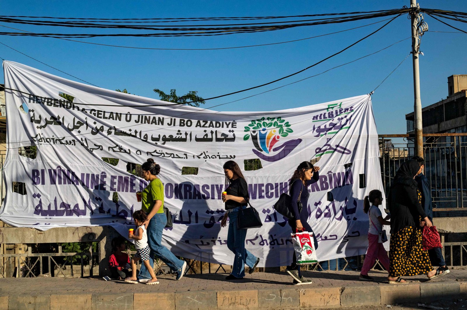 Syrian Kurdish women walk past a campaign banner for the PKK/YPG&#039;s so-called elections in the northeastern city of Qamishli, Syria, June 6, 2024. (AFP Photo)