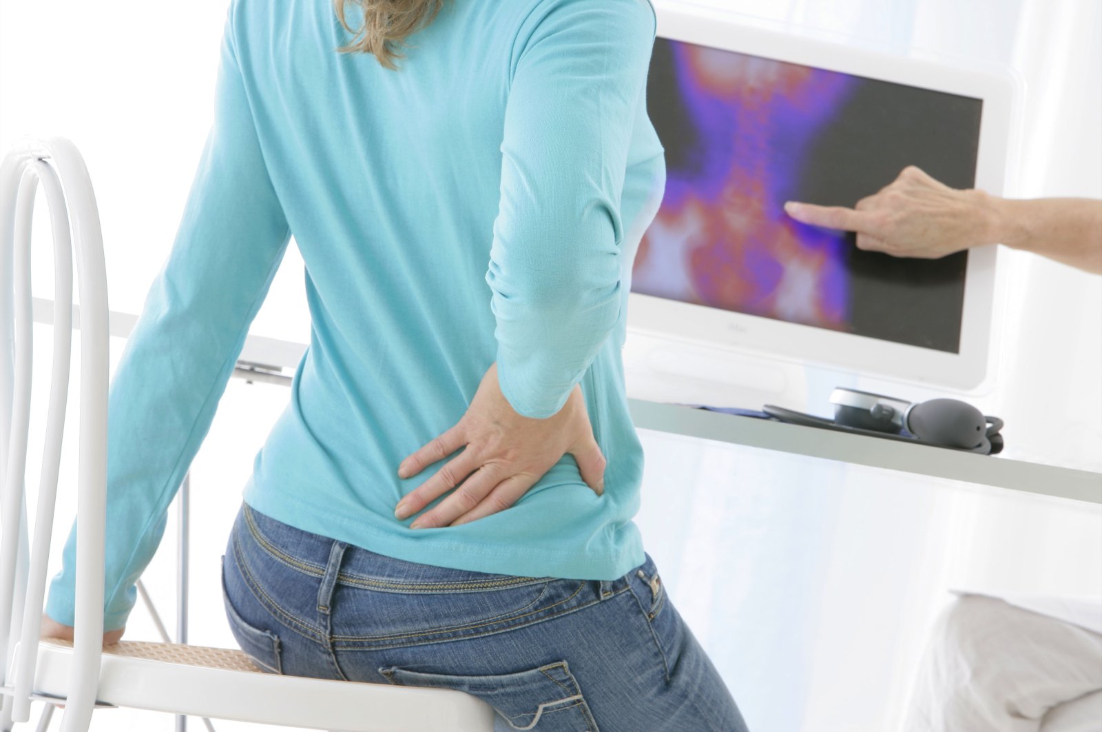 A woman is holding her back due to back pain, with an x-ray of the spinal column visible on the screen. (Getty Images)