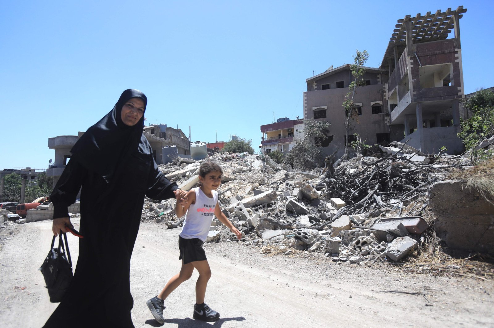 A woman holds the hand of a girl as they walk past buildings destroyed during previous Israeli military fire on the southern Lebanese village of Aita al-Shaab, near the border with northern Israel on June 29, 2024. (AFP Photo)