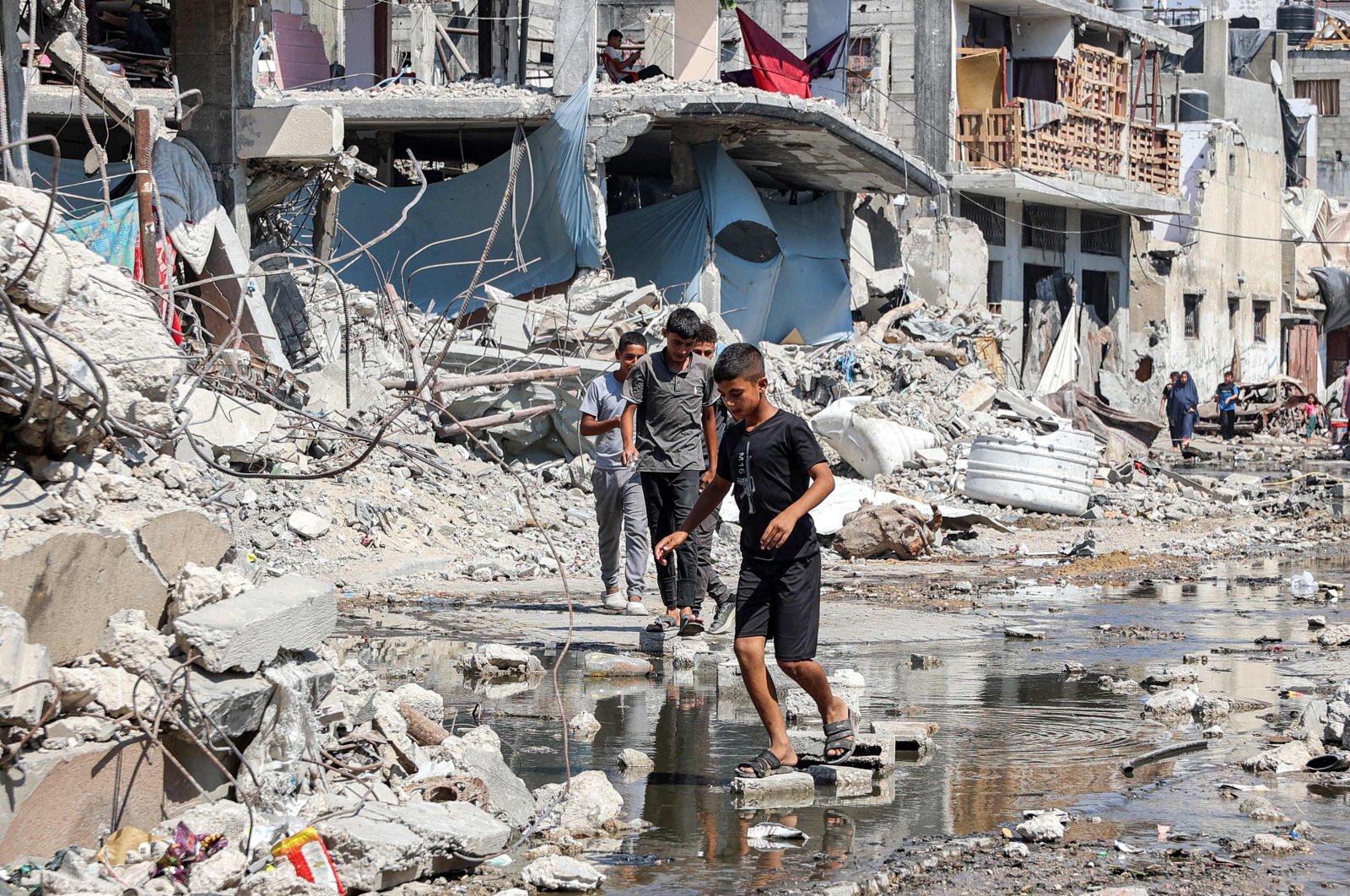 Boys walk on dry bricks to traverse a puddle of sewage water past mounds of trash and rubble along a street in the Jabalia camp for Palestinian refugees after Israel&#039;s destructive attacks in the northern Gaza Strip, Aug.14, 2024 (AFP Photo)