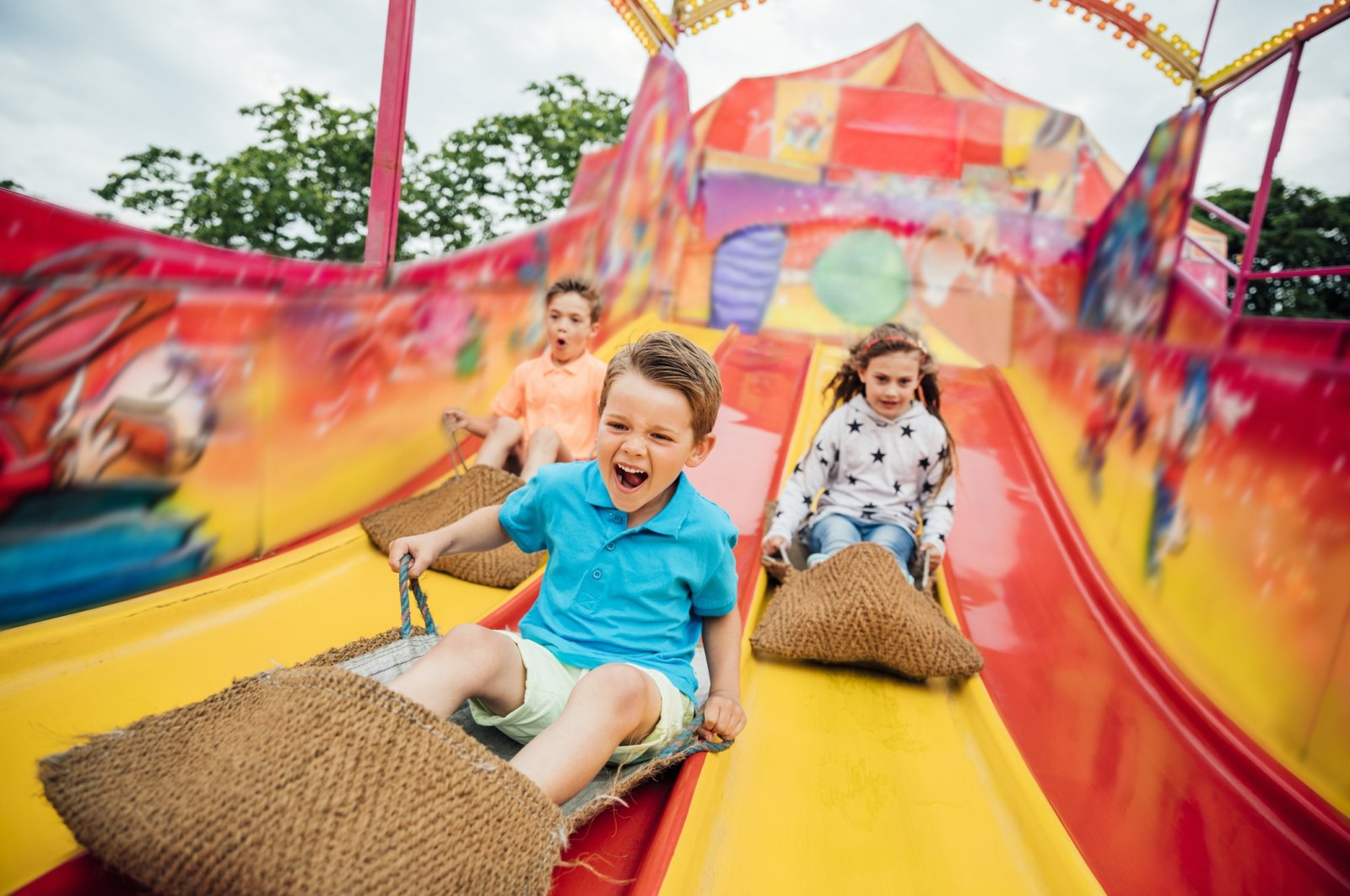 Children having fun sliding down a yellow and red slide while sitting in burlap sacks. (Getty Images Photo)