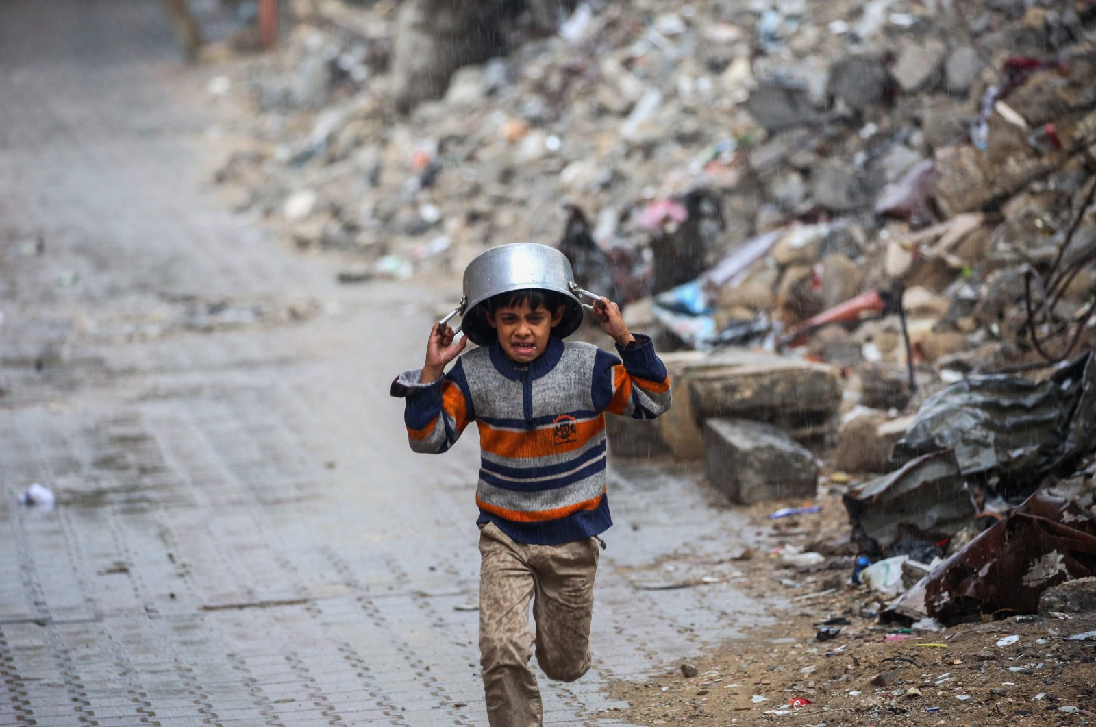 A displaced boy covers his head with a pan as he runs from the rain past buildings destroyed in Israeli attacks on the Bureij refugee camp, central Gaza Strip, Palestine, Nov. 24, 2024. (AFP Photo)