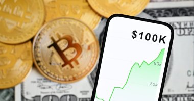 This illustration shows a mobile screen with a $100K mark, a representation of the Bitcoin cryptocurrency and U.S. dollar banknotes, Dec. 5, 2024. (AA Photo)