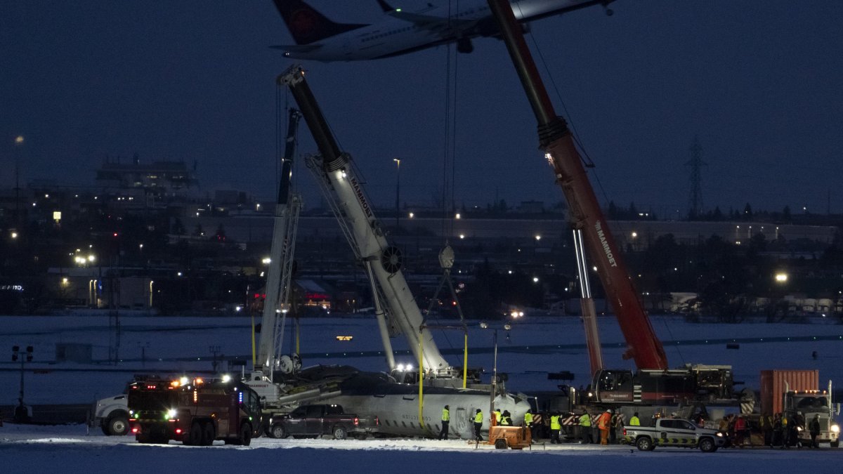 An Air Canada flight takes off in the background as cranes position to remove the wreckage of Delta flight 4819 from the runway at Toronto Pearson International Airport, Mississauga, Ontario, Canada, Feb. 19, 2025. (AP Photo)