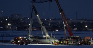 An Air Canada flight takes off in the background as cranes position to remove the wreckage of Delta flight 4819 from the runway at Toronto Pearson International Airport, Mississauga, Ontario, Canada, Feb. 19, 2025. (AP Photo)