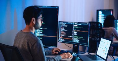 A software developer works on multiple screens displaying code and application diagrams. (Shutterstock File Photo)