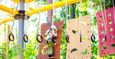 Gymnastic rings hang from ropes in a workout area as a young boy navigates a climbing panel in the background at an amusement park. (Shutterstock Photo) 