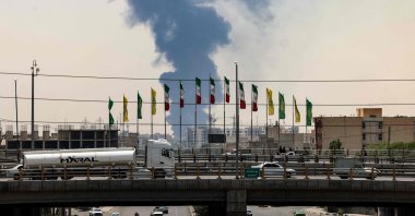 Flags flutter along a bridge as a plume of heavy smoke and fire rise from an oil refinery in southern Tehran, after it was hit in an overnight Israeli strike, June 15, 2025. (AFP Photo)