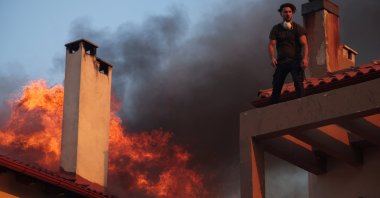 A local stands on a rooftop as flames rise from a burning house, while a wildfire burns in the village of Kryoneri, near Athens, Greece, July 26, 2025. (Reuters Photo)