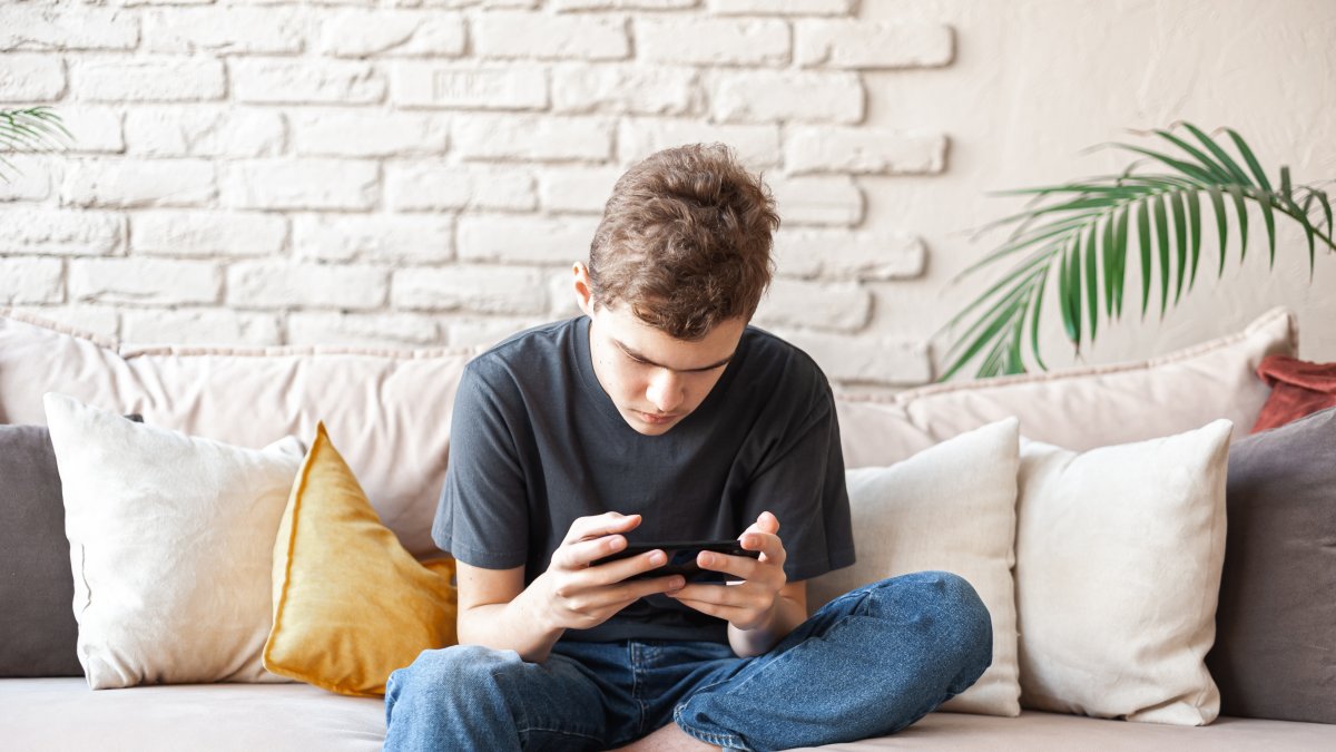A teenage boy sits on a couch, focused on his smartphone while playing games. (Shutterstock Photo) 