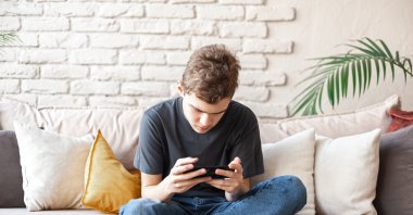 A teenage boy sits on a couch, focused on his smartphone while playing games. (Shutterstock Photo) 