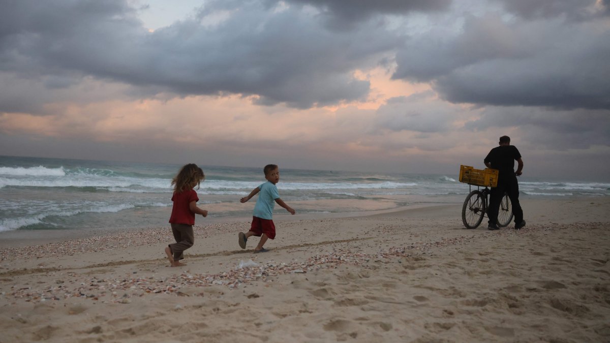 Displaced Palestinian children run behind a man pushing his bicycle on the sand at sunrise at a makeshift camp by the beach in Al-Zawayda city, near Deir al-Balah, in the central Gaza Strip on October 9, 2025, following an overnight announcement of a ceasefire deal between Hamas and Israel to be signed in Egypt. (Photo by Bashar TALEB / AFP)