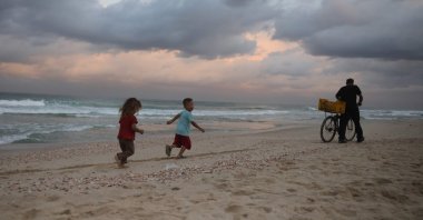 Displaced Palestinian children run behind a man pushing his bicycle on the sand at sunrise at a makeshift camp by the beach in Al-Zawayda city, near Deir al-Balah, in the central Gaza Strip on October 9, 2025, following an overnight announcement of a ceasefire deal between Hamas and Israel to be signed in Egypt. (Photo by Bashar TALEB / AFP)