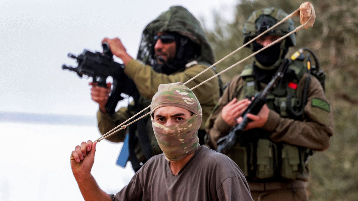 Israeli army soldiers stand behind a masked man swinging a slingshot while hurling stones at Palestinians who had gathered for the annual olive harvest season, during an attack by Israeli settlers on the Palestinian village of Beita, south of Nablus in the occupied West Bank, on Oct. 10, 2025. (AFP Photo)