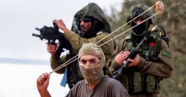 Israeli army soldiers stand behind a masked man swinging a slingshot while hurling stones at Palestinians who had gathered for the annual olive harvest season, during an attack by Israeli settlers on the Palestinian village of Beita, south of Nablus in the occupied West Bank, on Oct. 10, 2025. (AFP Photo)