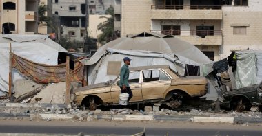 A Palestinian man carries containers past makeshift shelters and a destroyed car along a road in the Gaza Strip, Palestine, Nov. 9, 2025. (AFP Photo)
