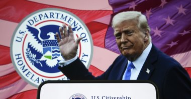 This illustration photo shows a logo of the U.S. Citizenship and Immigration Services on a smartphone with President Donald Trump and the logo of the Homeland Security Department in the background, Dec. 19, 2025. (AA Photo)