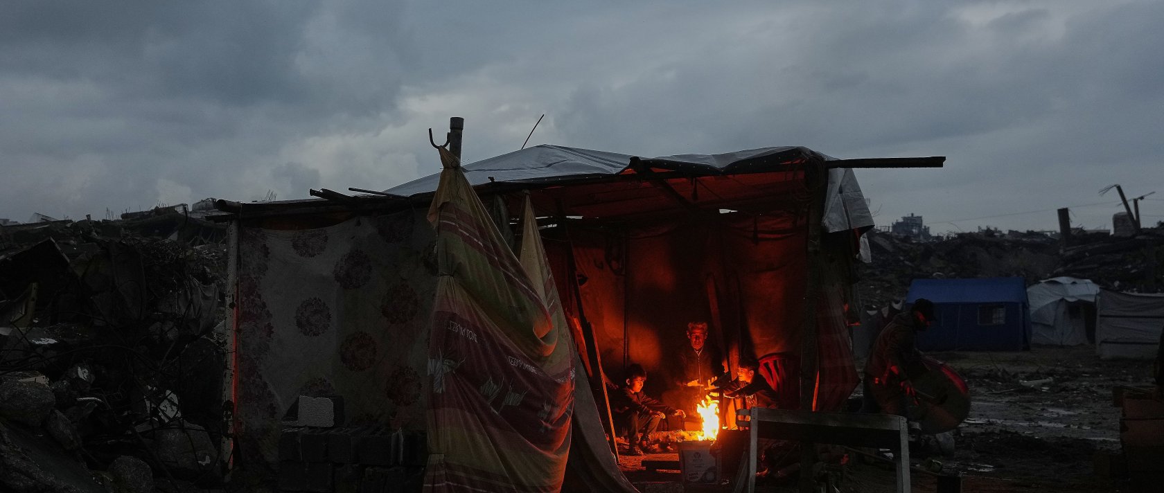 Aziz Halawa, 60, sits with his children as they warm themselves next to the fire inside their tent amid the destruction left by the Israeli air and ground operations, Gaza City, Palestine, Dec. 12, 2025. (AP Photo)
