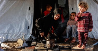 A displaced Palestinian woman heats a pot as children look on outside her tent-home as the region experiences rain and cold winter conditions, in Gaza City, Palestine, Dec. 28, 2025. (AFP Photo)