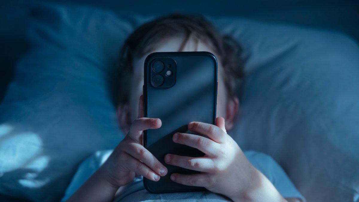 A child lies in bed at night, holding a mobile phone close to his face. (Shutterstock Photo)