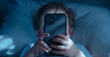 A child lies in bed at night, holding a mobile phone close to his face. (Shutterstock Photo)