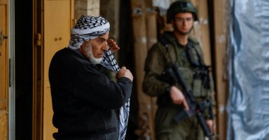 A Palestinian man puts on a keffiyah as an Israeli soldier stands guard in the old city in Hebron in the Israeli-occupied West Bank, Feb. 9, 2026. (Reuters Photo)