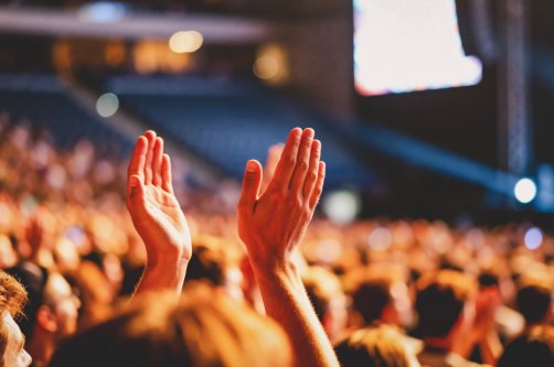 A close-up view from within a large crowd shows two hands raised high, clapping together in appreciation during a dimly lit indoor performance or presentation. (Shutterstock Photo)