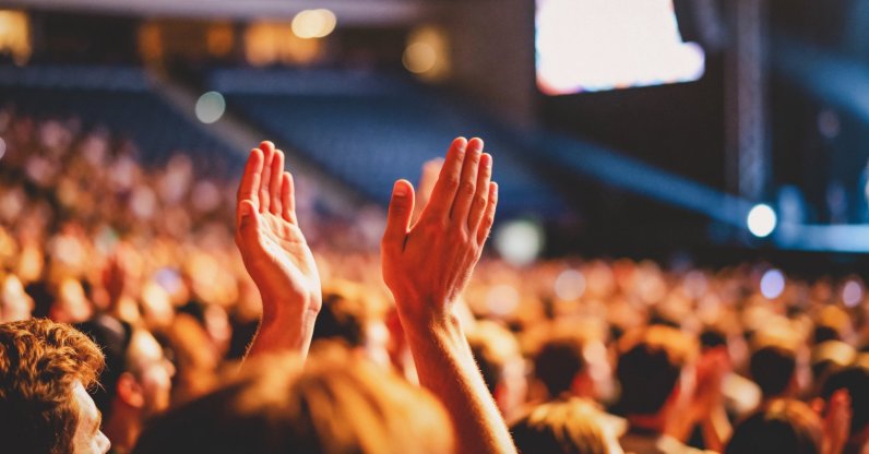 A close-up view from within a large crowd shows two hands raised high, clapping together in appreciation during a dimly lit indoor performance or presentation. (Shutterstock Photo)