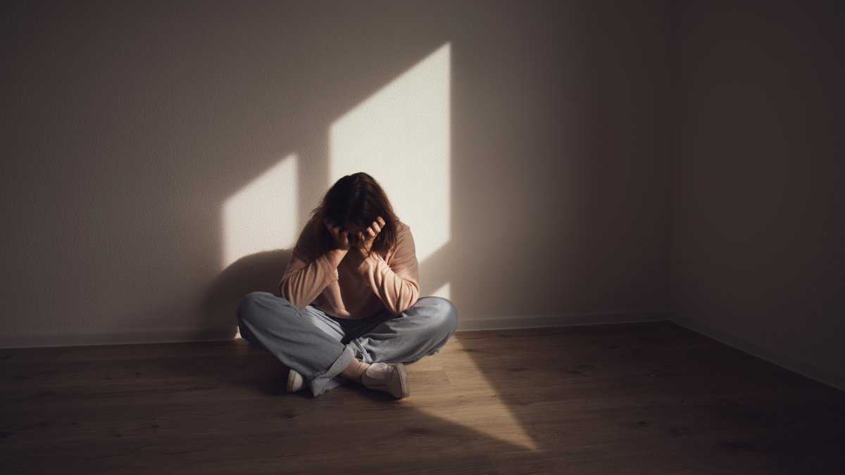 A young woman sits on the floor in a dark room, holding her head in anguish. (Shutterstock Photo)