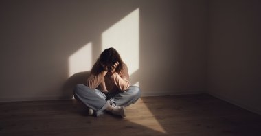 A young woman sits on the floor in a dark room, holding her head in anguish. (Shutterstock Photo)