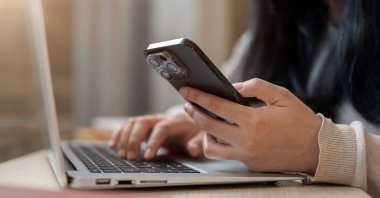 A close-up image of a woman using her smartphone while working on her laptop. (Shutterstock Photo)