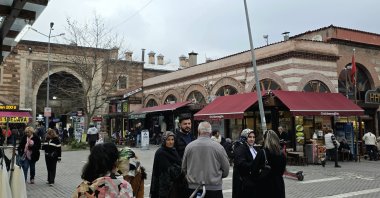 People walk past a historic stone building with arched entrances and shops, Bursa, Türkiye, March 23, 2026. (Shutterstock Photo)