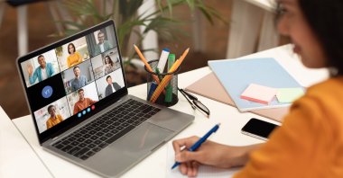 A woman sits at a desk with a laptop open, displaying a video conference meeting with several participants. (Shutterstock Photo)