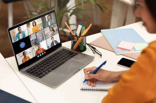 A woman sits at a desk with a laptop open, displaying a video conference meeting with several participants. (Shutterstock Photo)