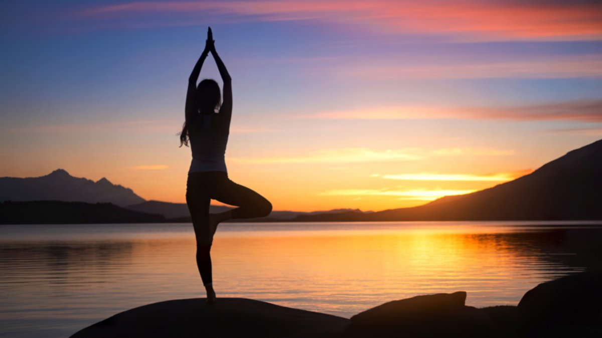 A woman stands in a tree pose, yoga position, on a rock by a lake, silhouetted against a vibrant sunset sky and mountain range. (Shutterstock Photo)