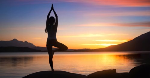 A woman stands in a tree pose, yoga position, on a rock by a lake, silhouetted against a vibrant sunset sky and mountain range. (Shutterstock Photo)